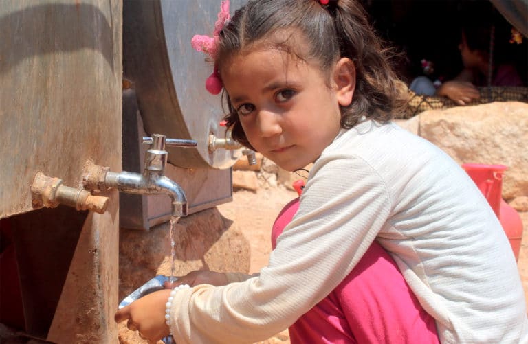 Jente fra Syria som sitter på huk og ser i kameraet mens hun tapper vann fra en kran i en flyktningleir.