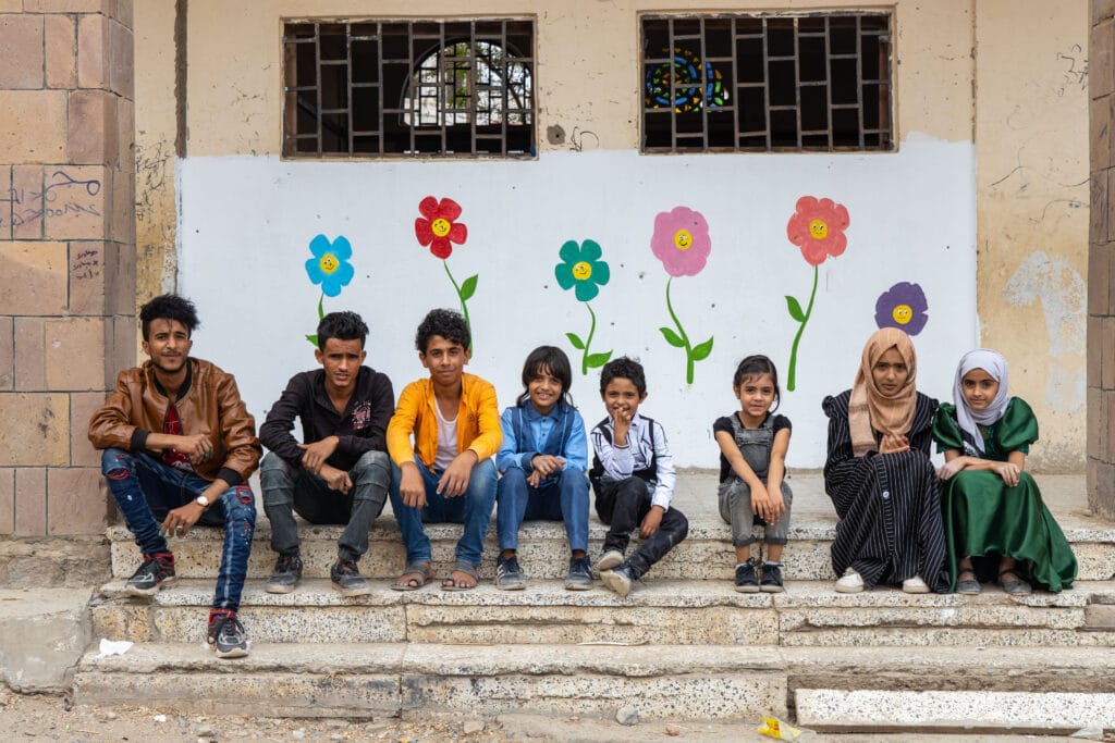 En gruppe barn i alle aldre sitter på en trapp foran en ødelagt skole, hvor de har malt blomster på veggen.
