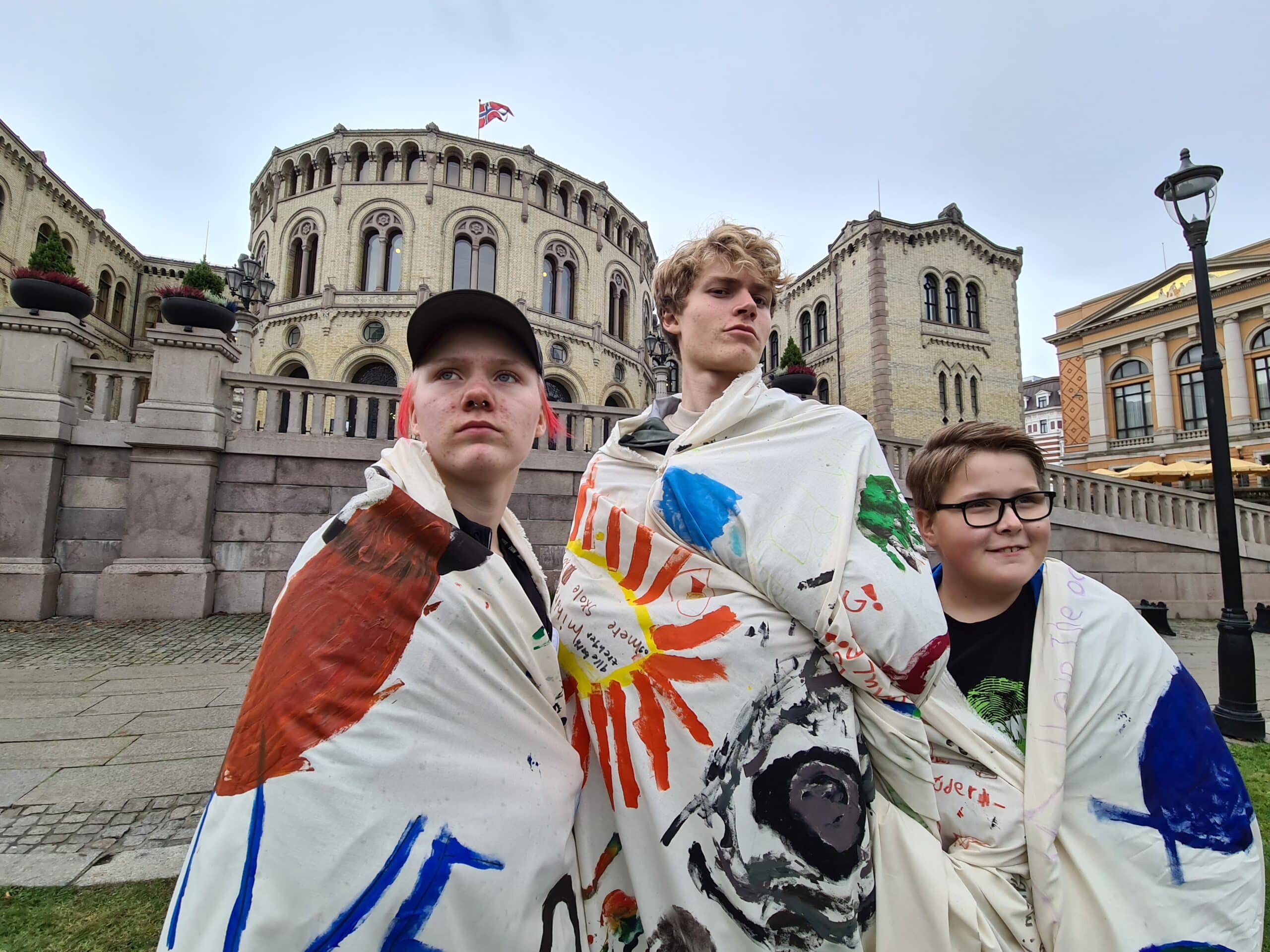 3 ungdommer står utenfor Stortinget og poserer med klimaparoler som de har pakket seg inn i