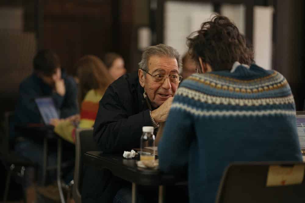En eldre mann med briller sitter dypt engasjert i en samtale med en yngre person.