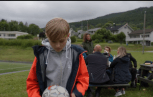 Fotografi av en gutt som holder en fotball og ser ned. Bak han sitter en gruppe foreldre