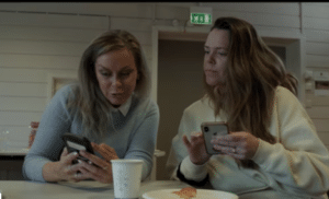 Fotografi av to damer som sitter ved et bord og titter ned i mobiltelefonen