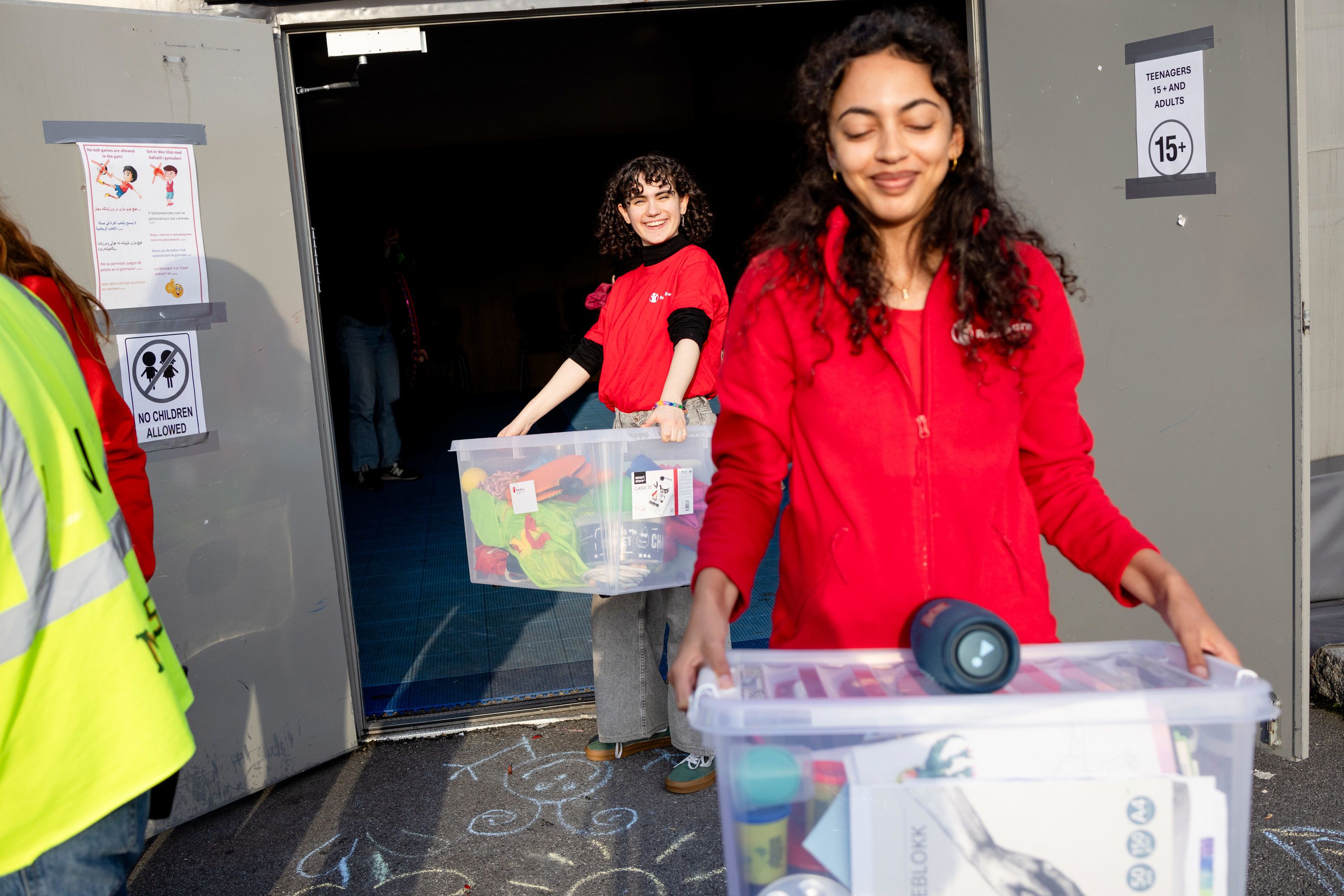 Sara og Amanda i røde Redd Barna gensere går og bærer på store plastbokser med leker. 