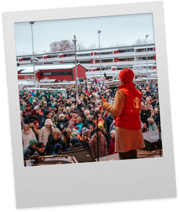 Polaroidbilde av en dame med rød Redd Barna vest og rød lue som snakker til et stort publikum fra scenen. 