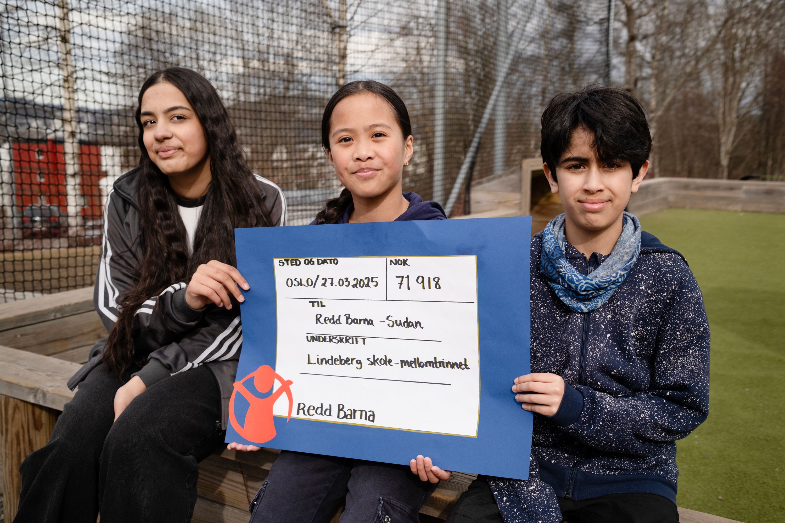 Mahnoor, Janine og og Adam sitter på uteplassen på Lindeberg skole og holder opp sjekken til Redd Barna. 