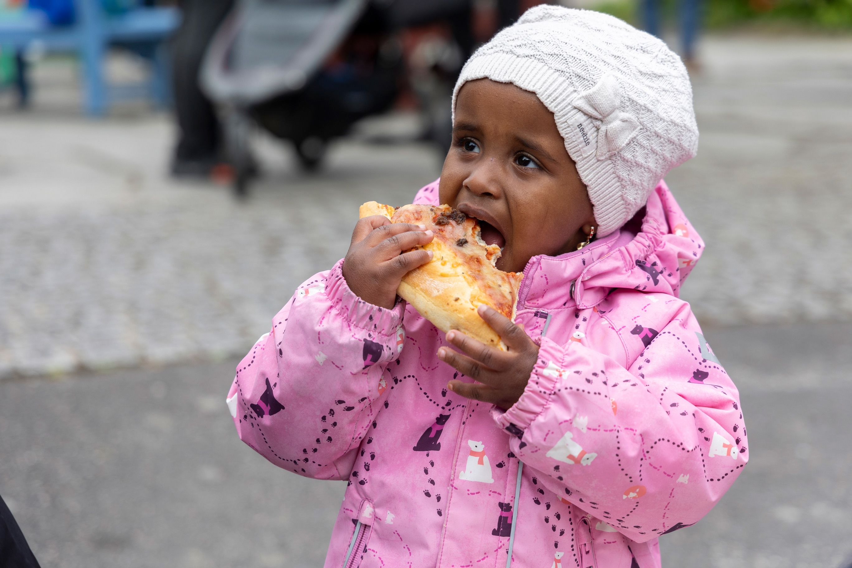 En liten jente med rosa jakke og hvit lue spiser pizza i Kristiansand Dyrepark.