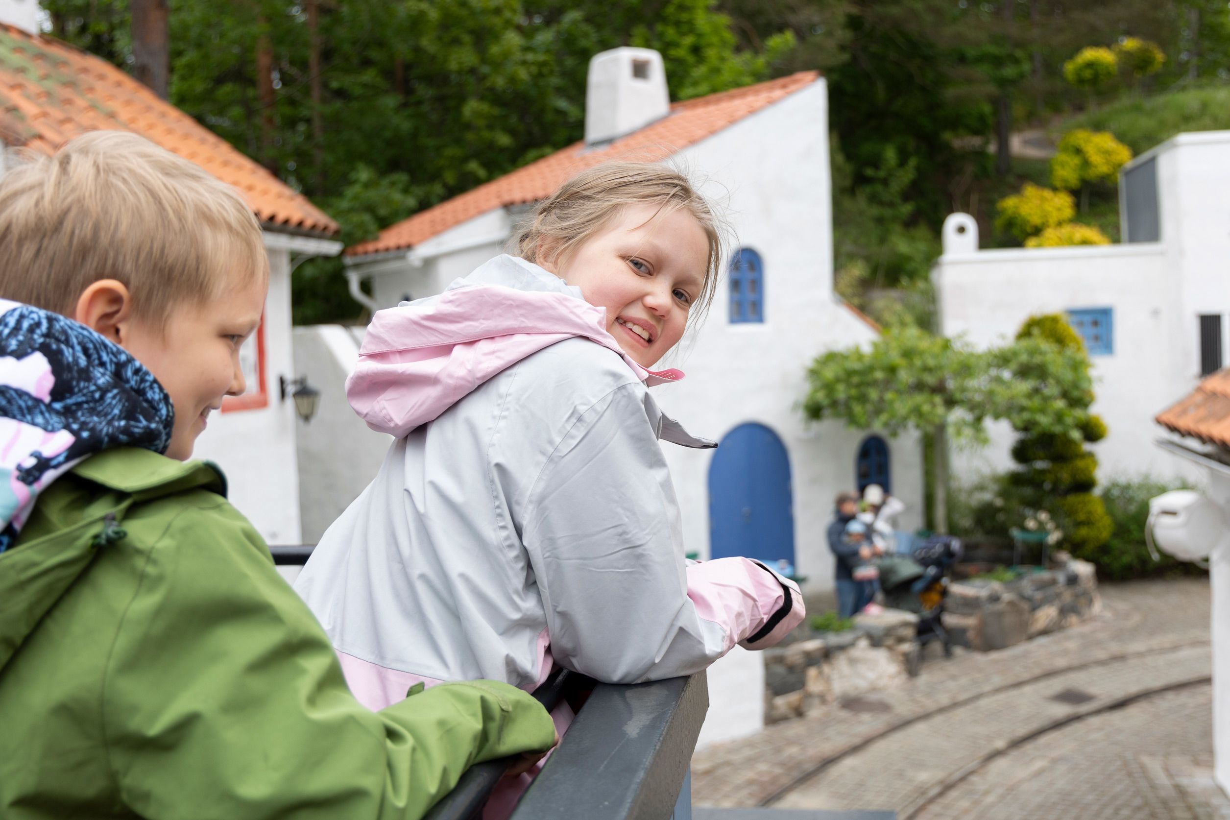 En jente med hvit jakke og lyst hår smiler til kamera med Kardemommeby i bakgrunnen.